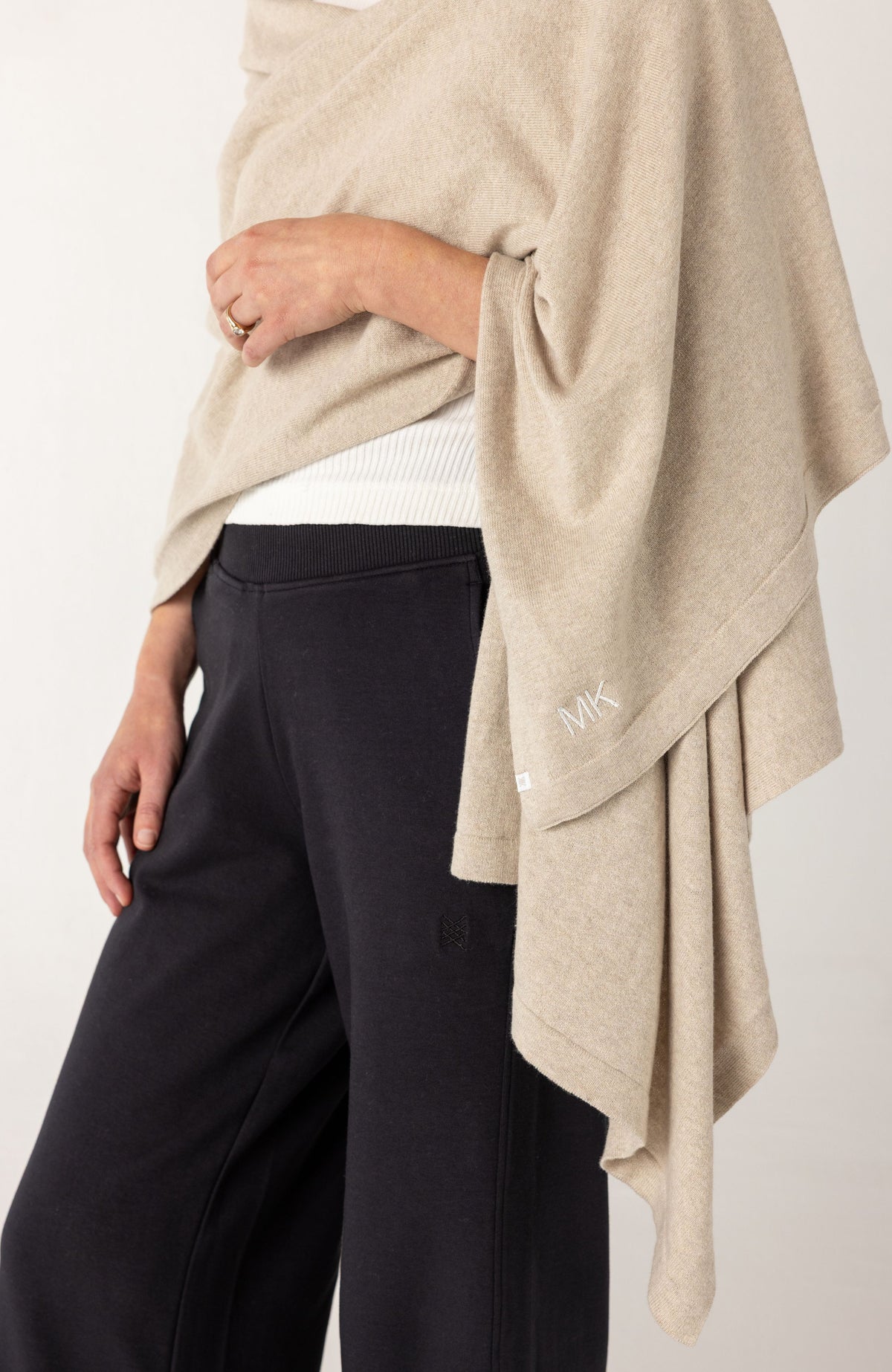 A close-up of a person wearing a draped beige knit poncho or shawl over a white top and black wide-leg pants, against a plain light background.
