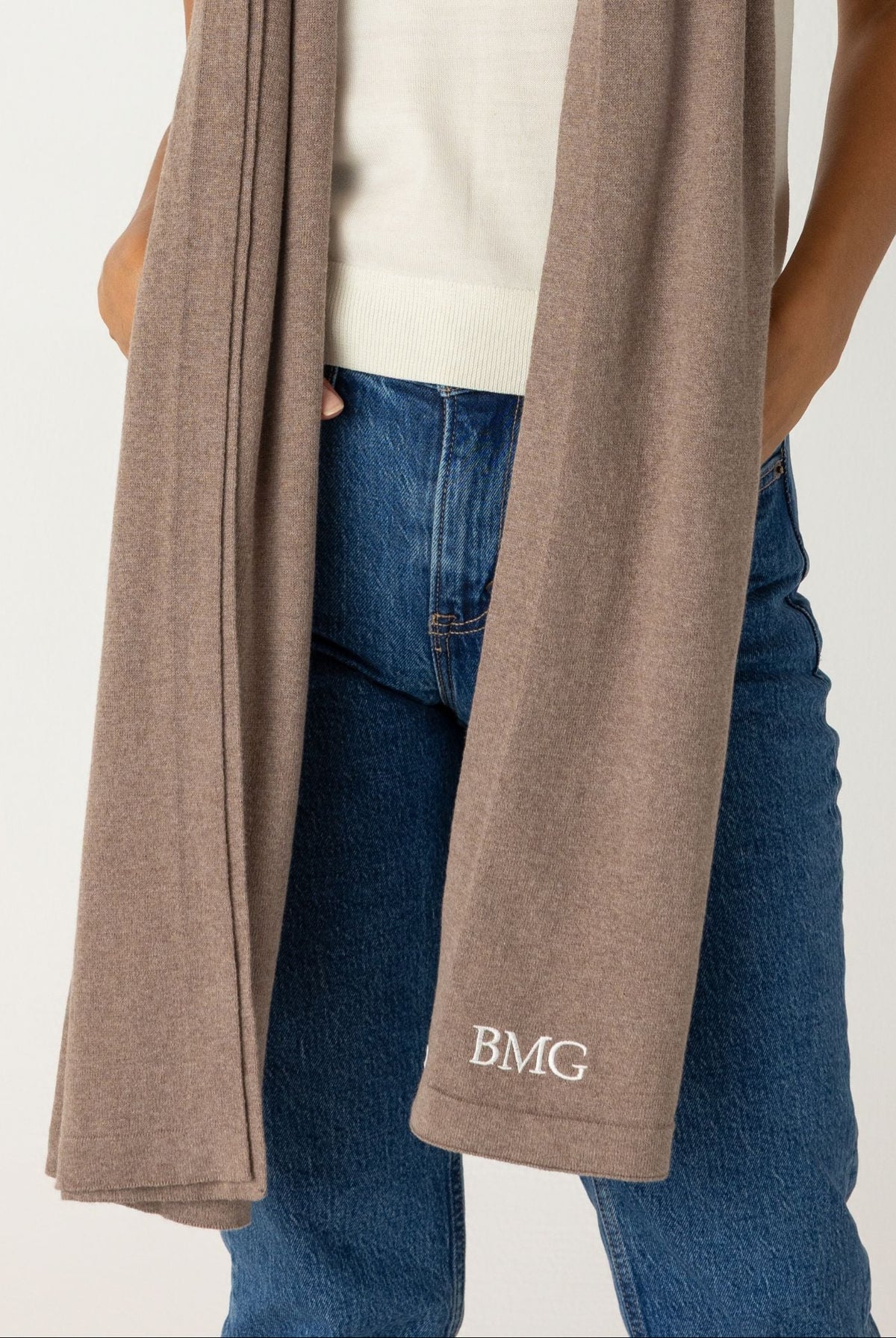 A woman modeling a taupe knit scarf with a white t-shirt and blue jeans, standing against a light gray studio background. The scarf features a small white embroidered logo 'BMG' near the bottom edge.