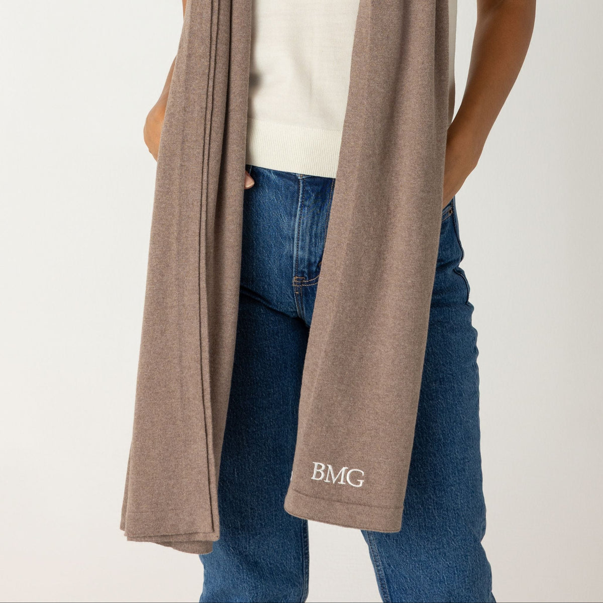 A woman modeling a taupe knit scarf with a white t-shirt and blue jeans, standing against a light gray studio background. The scarf features a small white embroidered logo 'BMG' near the bottom edge.