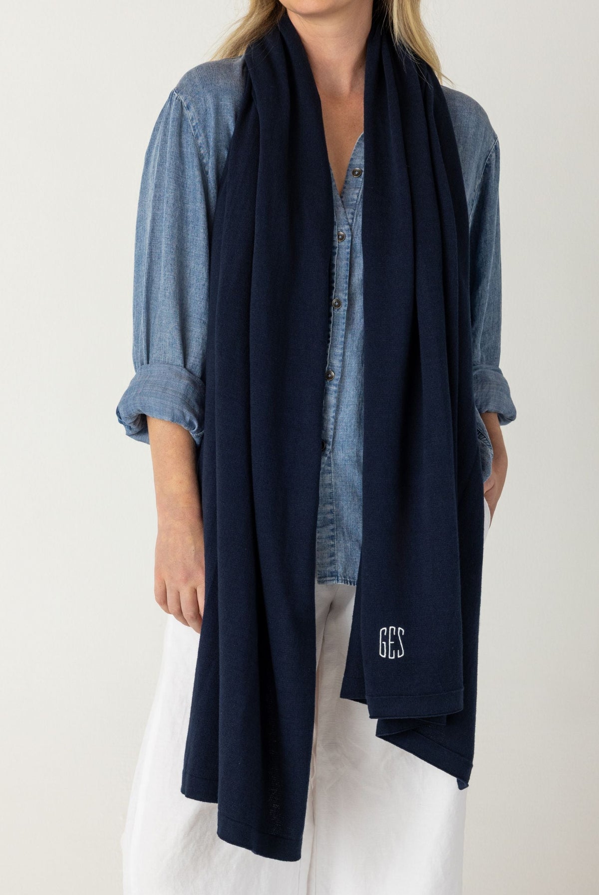 A woman modeling a dark navy blue knit scarf with a small white embroidered monogram, styled over a blue denim shirt and white pants against a plain light gray background.