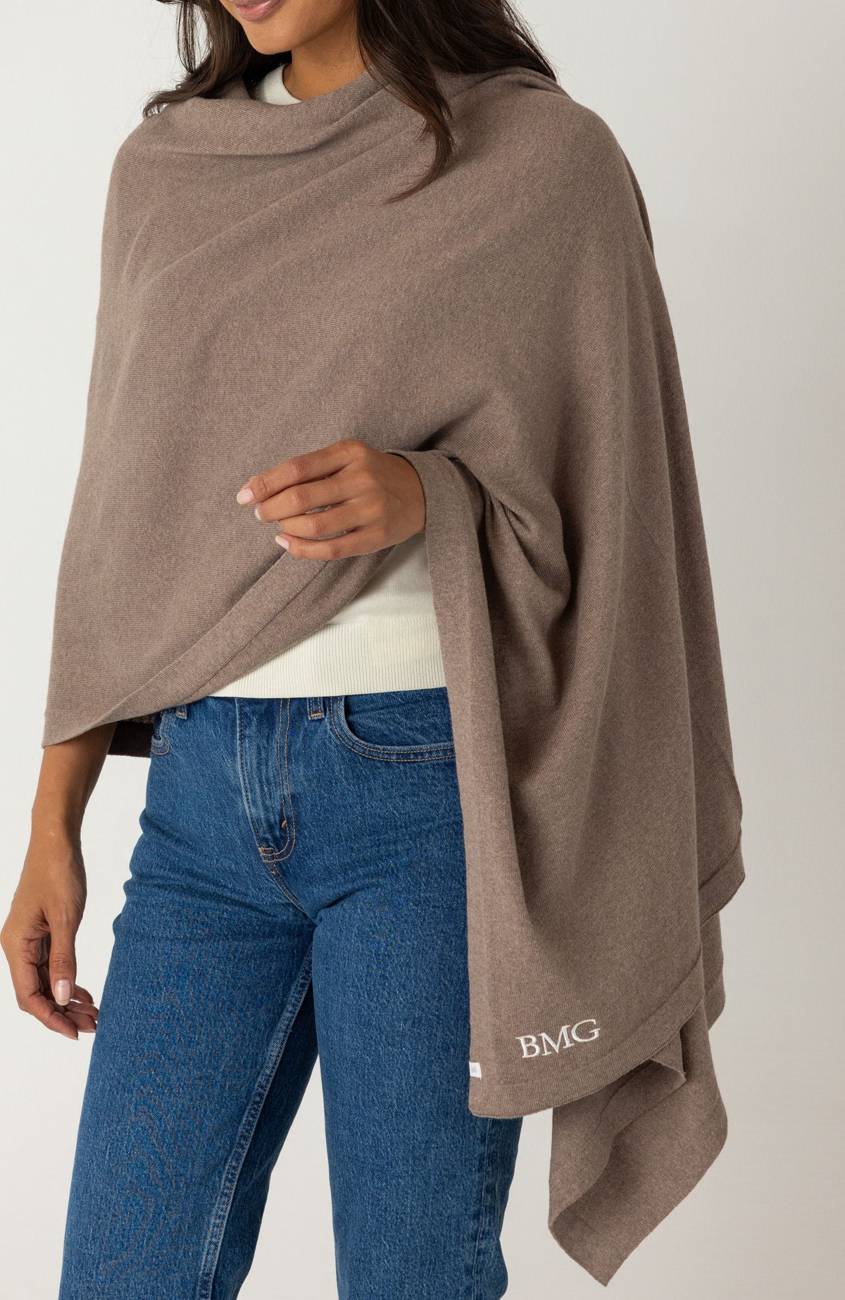 A woman wearing a taupe-brown draped wrap over a white top and blue jeans. The wrap has a small white embroidered 'BMG' logo near the lower right hem, shown against a plain light gray studio background.