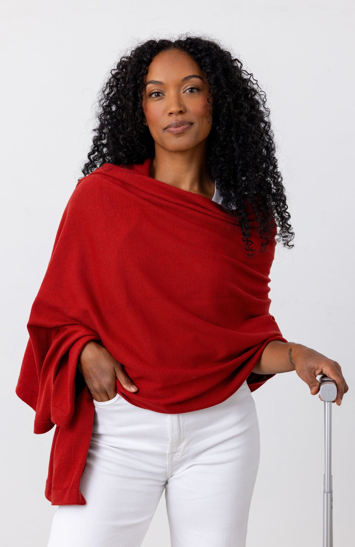 A woman with curly black hair wearing a bright red shawl draped over her shoulders and white pants, holding a silver suitcase handle against a plain white studio background.
