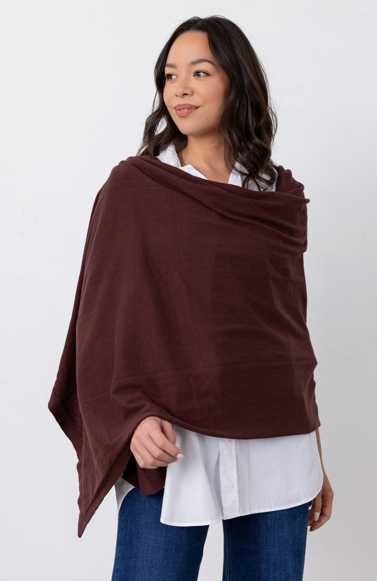 A woman wearing a dark brown shawl or poncho over a white button-down shirt and blue jeans, standing against a plain white studio background.