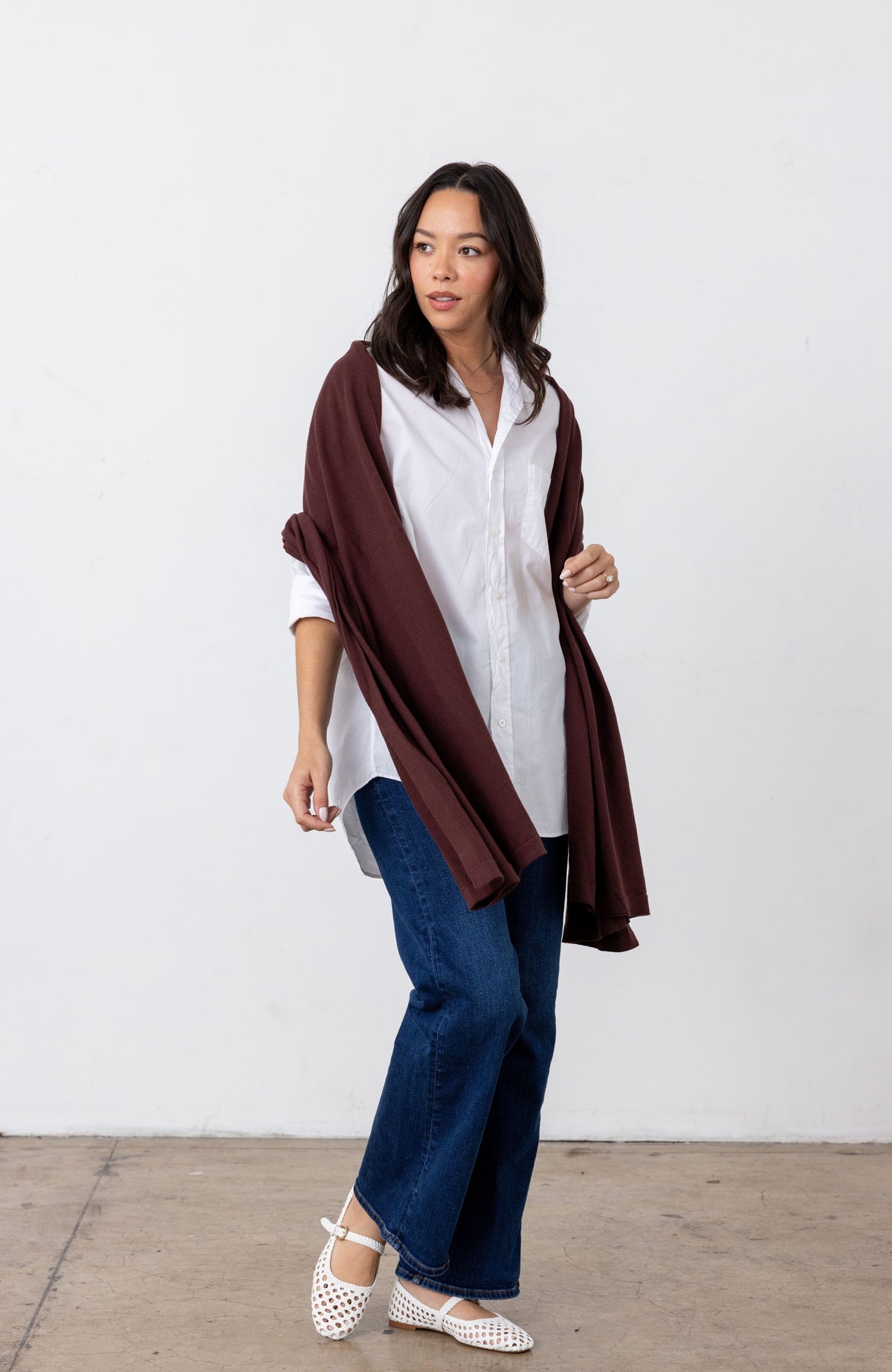 A woman modeling a dark brown shawl draped over a white button-down shirt, paired with blue jeans and white flat shoes, standing against a plain white wall.