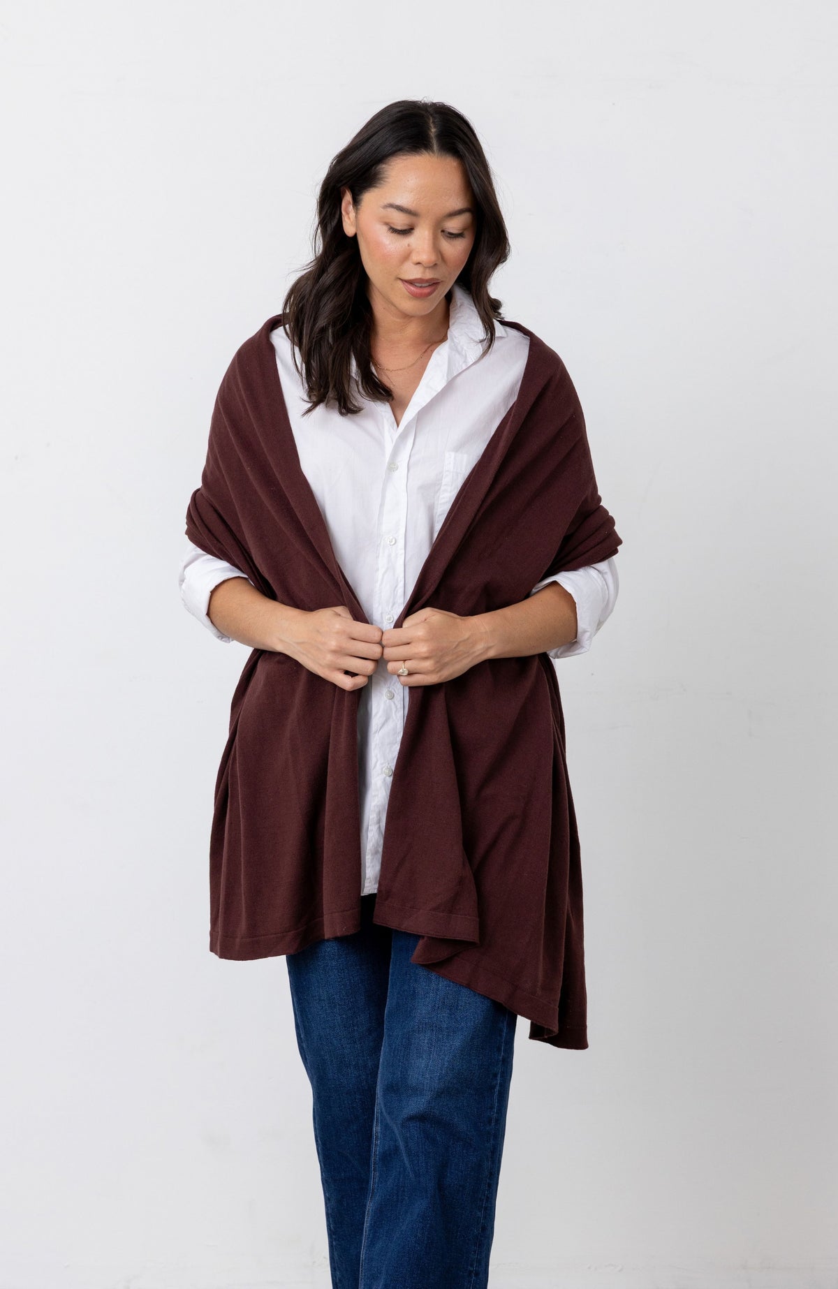 Woman in a white button down shirt and jeans wearing a dark reddish-brown scarf as a wrap around her shoulders against a white background