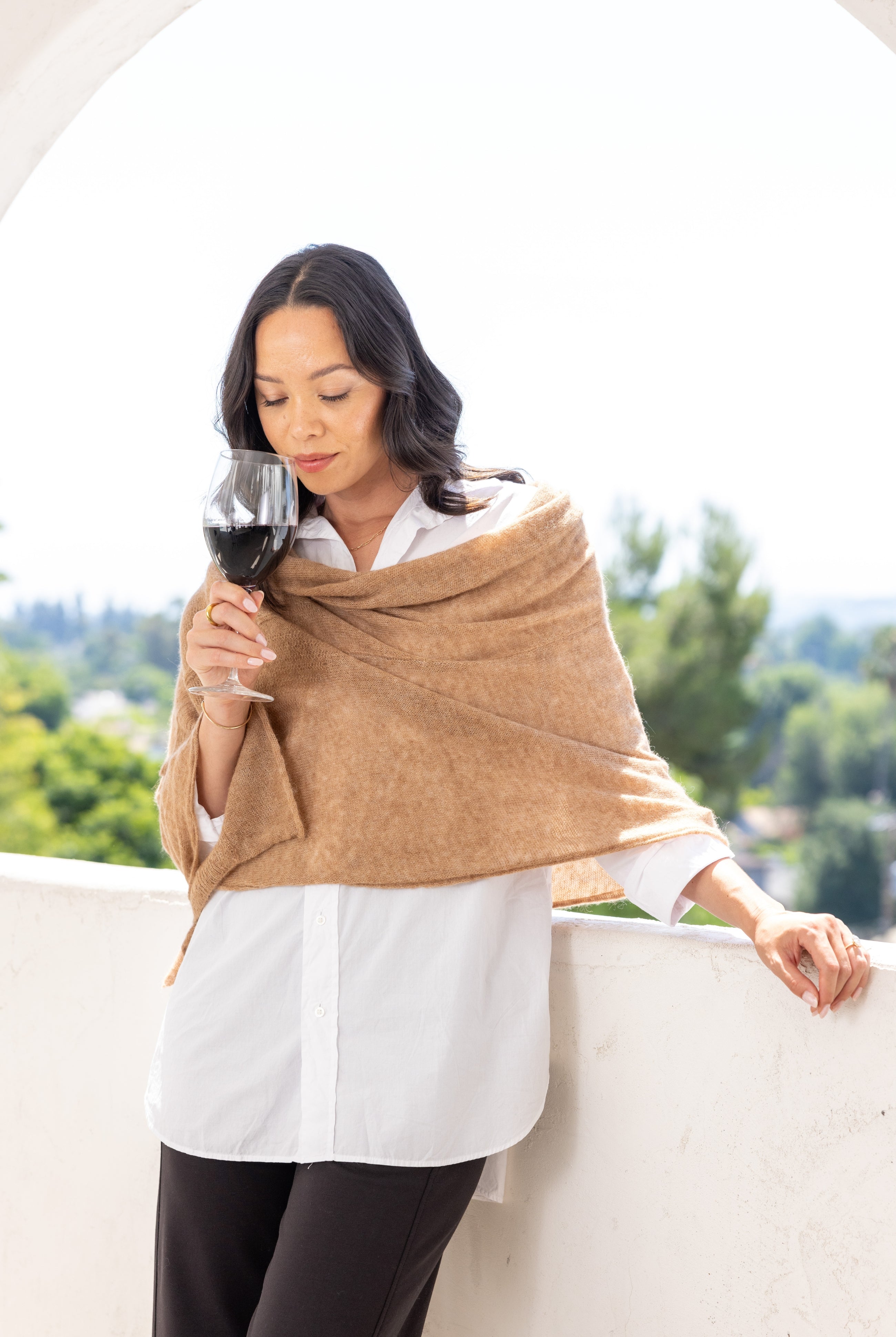 Woman shown wearing a beige scarf draped over her shoulders holding a glass of wine