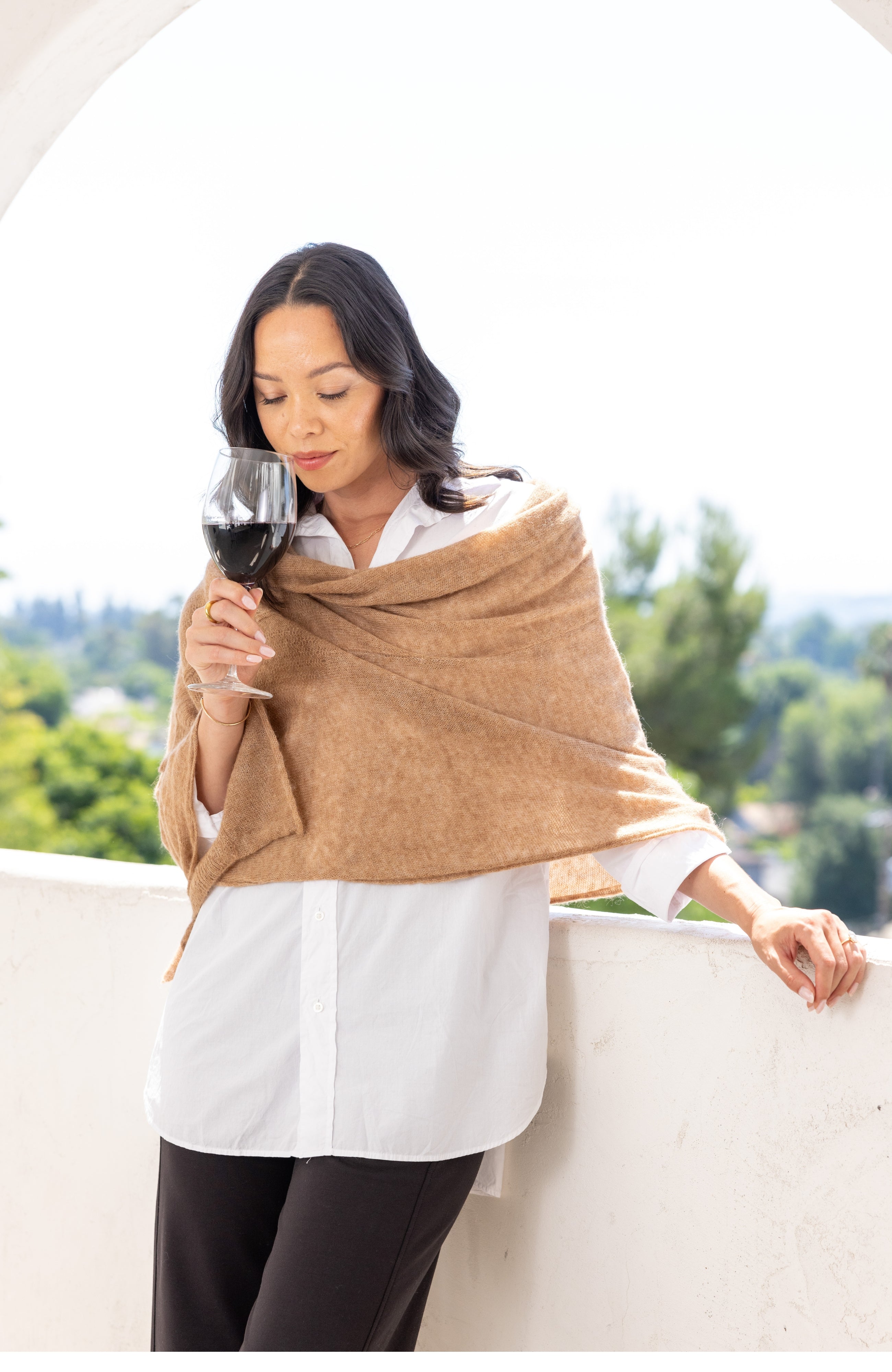 Woman shown wearing a beige scarf draped over her shoulders holding a glass of wine