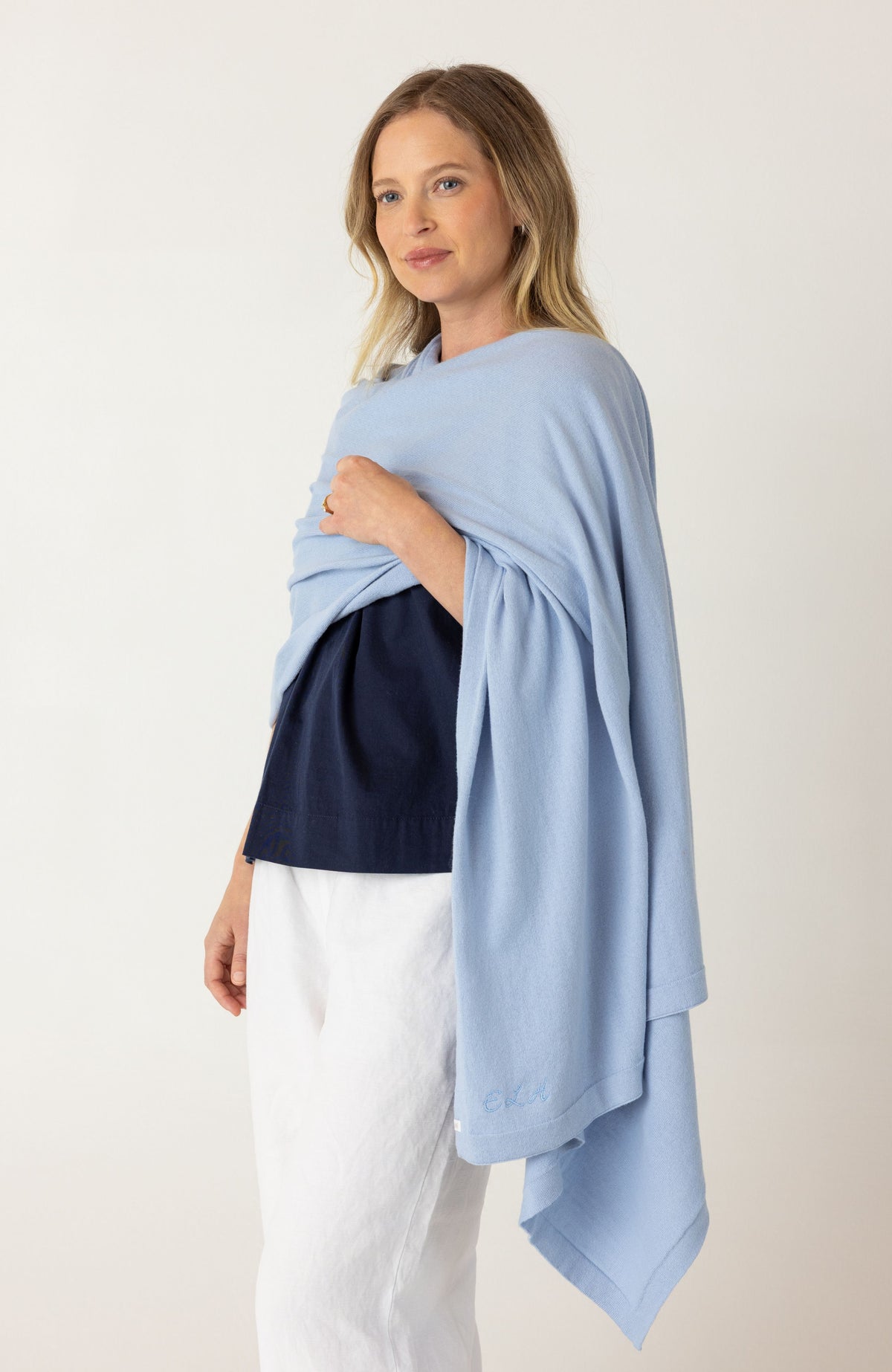 A woman modeling a light blue wrap or shawl draped over her shoulders, worn over a navy top and white pants, against a plain off-white studio background.