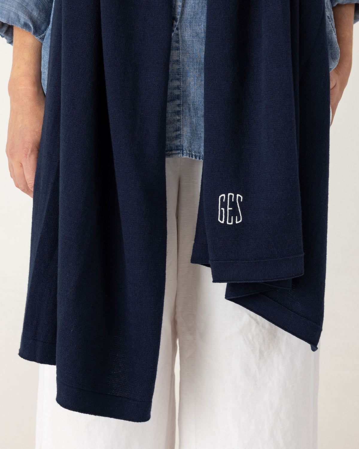 A navy blue scarf layered over a light blue denim shirt and white pants, with a small white embroidered monogram on the lower right hem. The photo is cropped from the neck down against a plain light background.