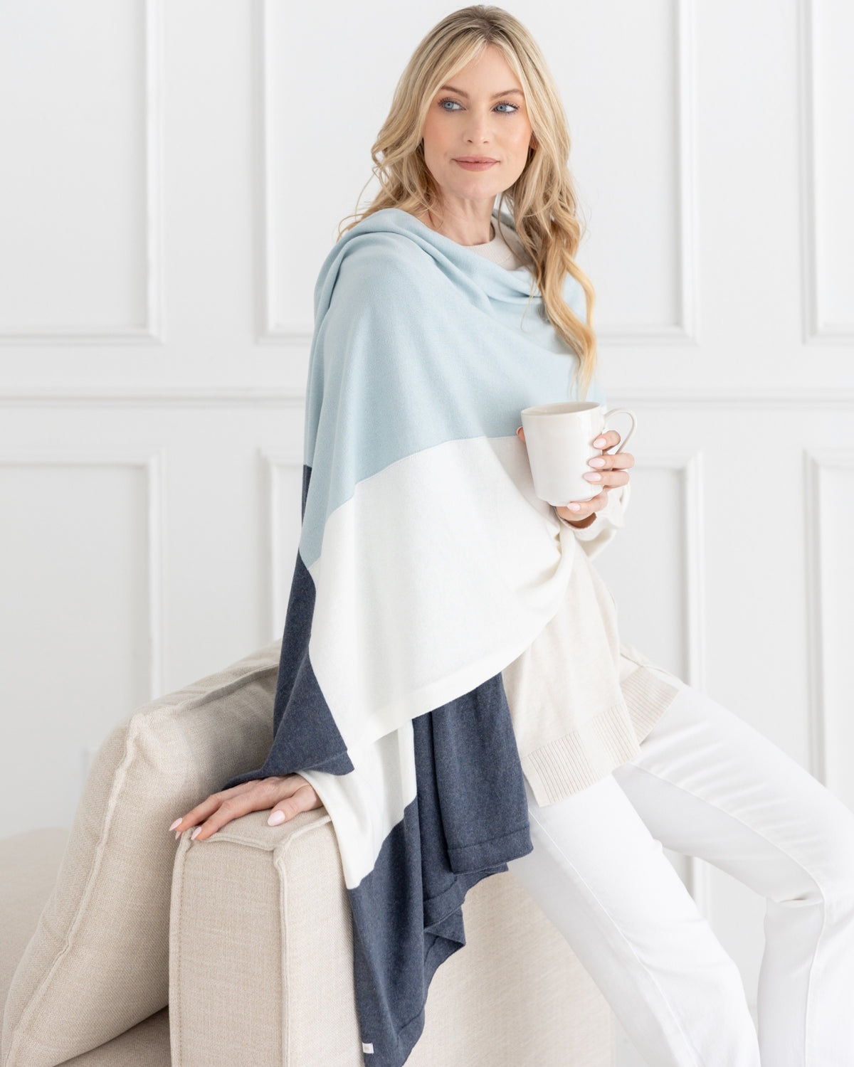 Woman leaning on side of sofa wearing the Dreamsoft Travel Scarf in Sky Blue Colorblock which is a light blue, gray and cream scarf, while holding her coffee mug