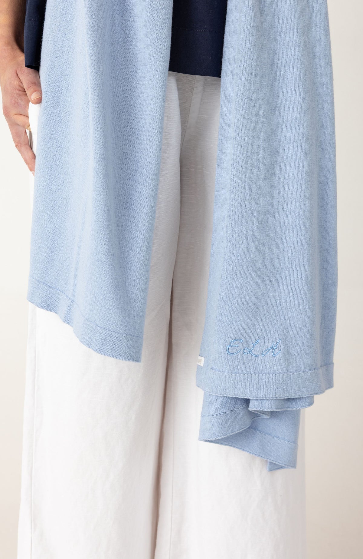 A close-up of a person wearing a light blue knit wrap draped over white wide-leg pants, with a dark navy top partially visible at the top. The cuff of the blue garment has a small embroidered script detail.