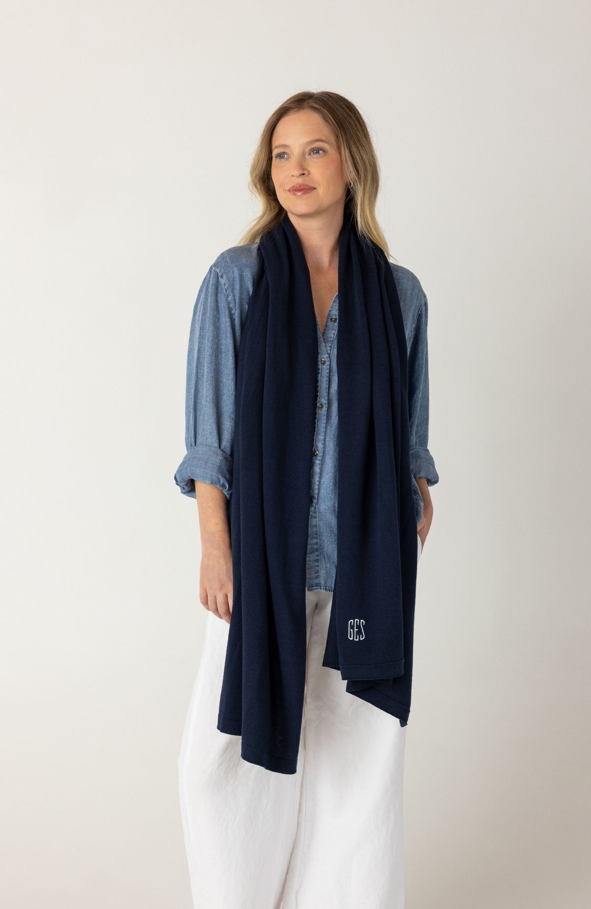 A woman modeling a dark navy blue knit scarf with a small white embroidered monogram, styled over a blue denim shirt and white pants against a plain light gray background.