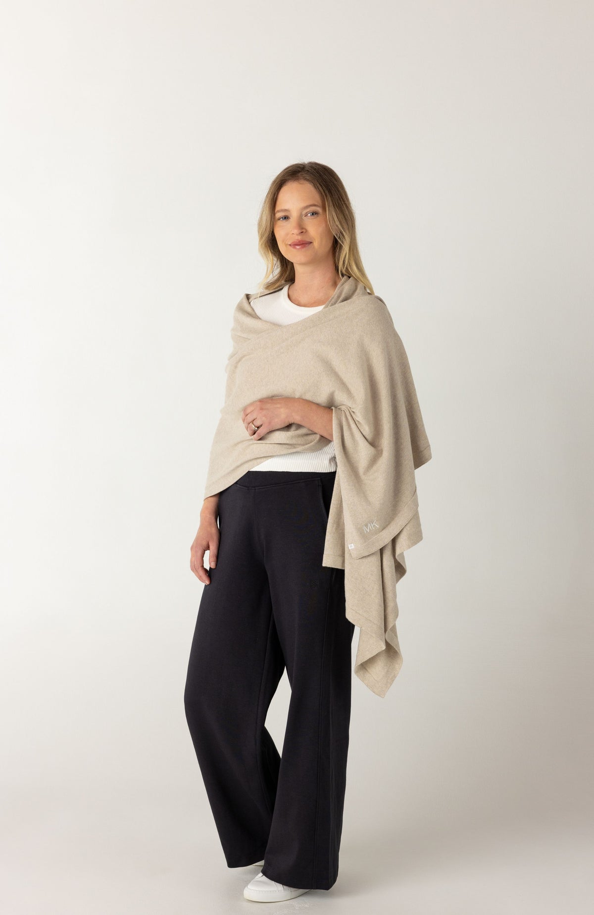 A woman modeling a light beige knit wrap or shawl draped over her shoulders, worn over a white top and paired with dark wide-leg pants, against a plain light gray studio background.