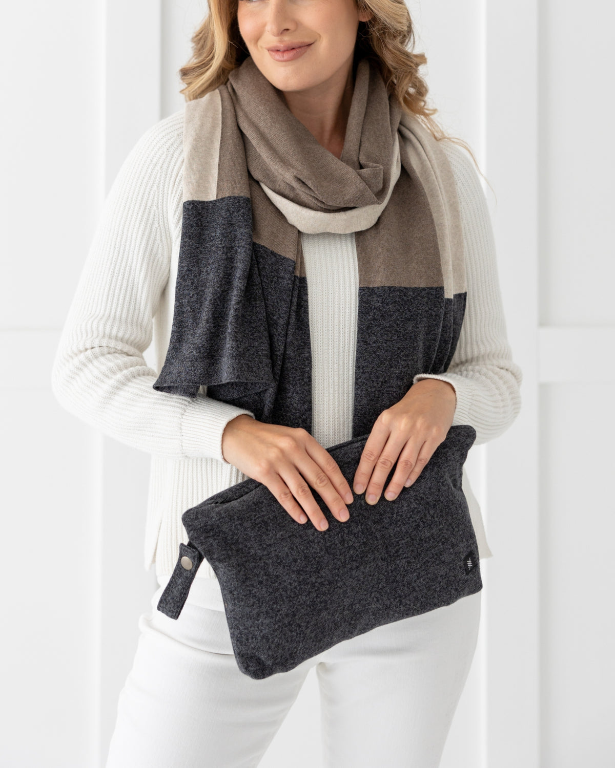 Woman holding Graphite Carry Pouch, which is a Gray zipper pouch that can hold the Dreamsoft Travel Scarf, woman is shown wearing a Brownstone Colorblock Dreamsoft Travel Scarf which is gray, beige and tan