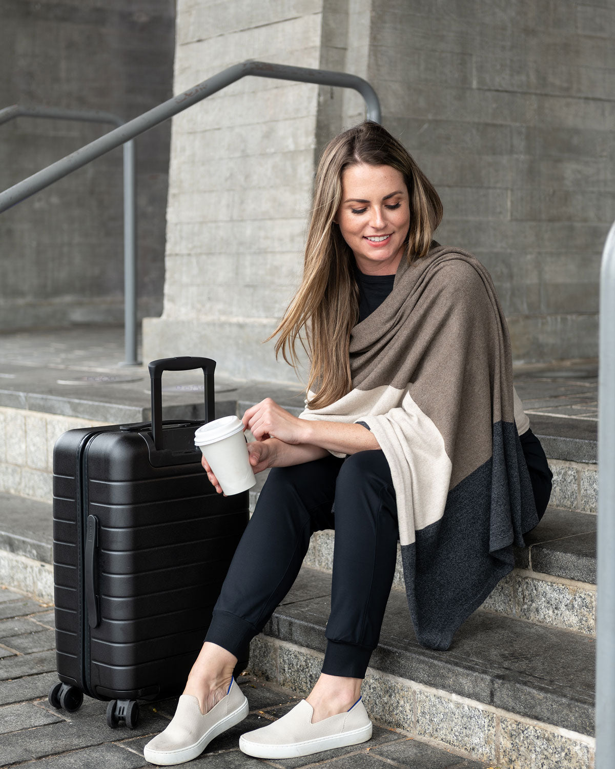 Woman wearing the Dreamsoft Travel Scarf in Brownstone Colorblock which is a brown, tan and gray scarf,  as a wrap while sitting on a step with her suitcase and coffee