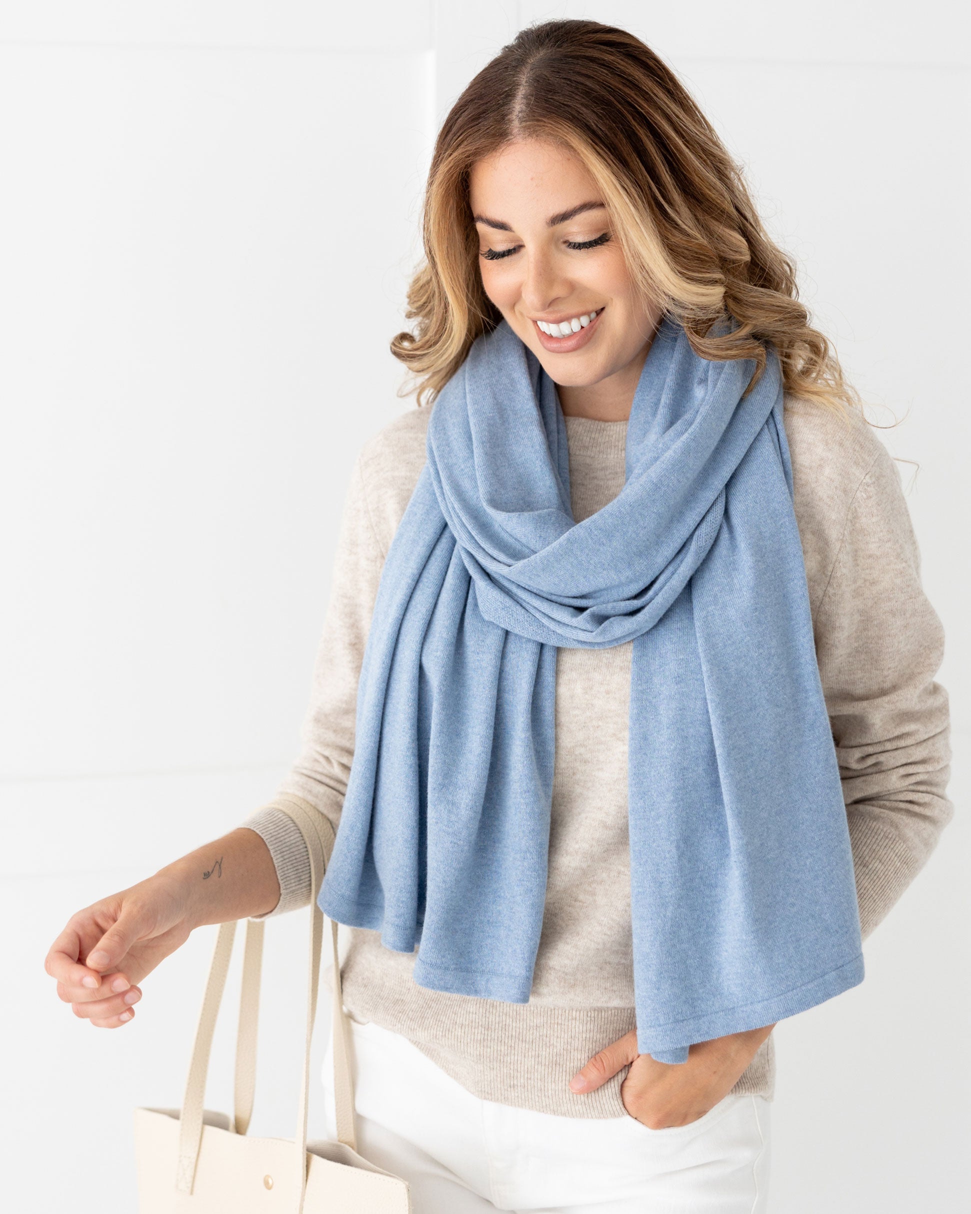 Woman wearing the Cashmere Cotton Luxe Travel Scarf in Horizon Blue which is a light blue scarf, pulling a suitcase while holding her coffee cup