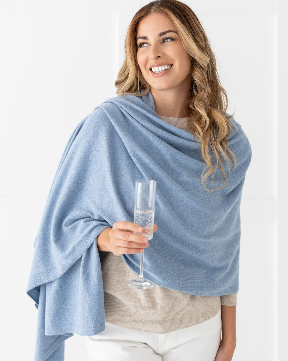 Woman wearing the Cashmere Cotton Luxe Travel Scarf in Horizon Blue which is a light blue scarf, with champagne glass in hand