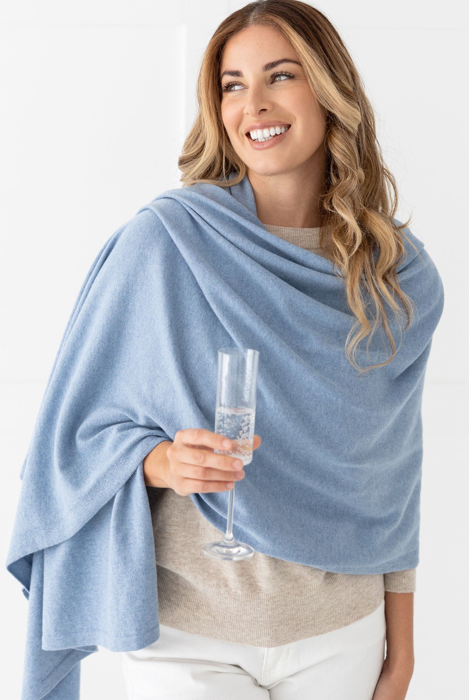 Woman wearing the Cashmere Cotton Luxe Travel Scarf in Horizon Blue which is a light blue scarf, with champagne glass in hand