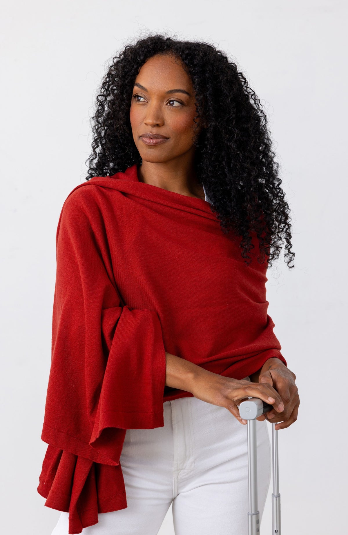 A woman with curly black hair wearing a draped red shawl or poncho-style top over a white top and white pants, holding a silver suitcase handle against a plain white studio background.