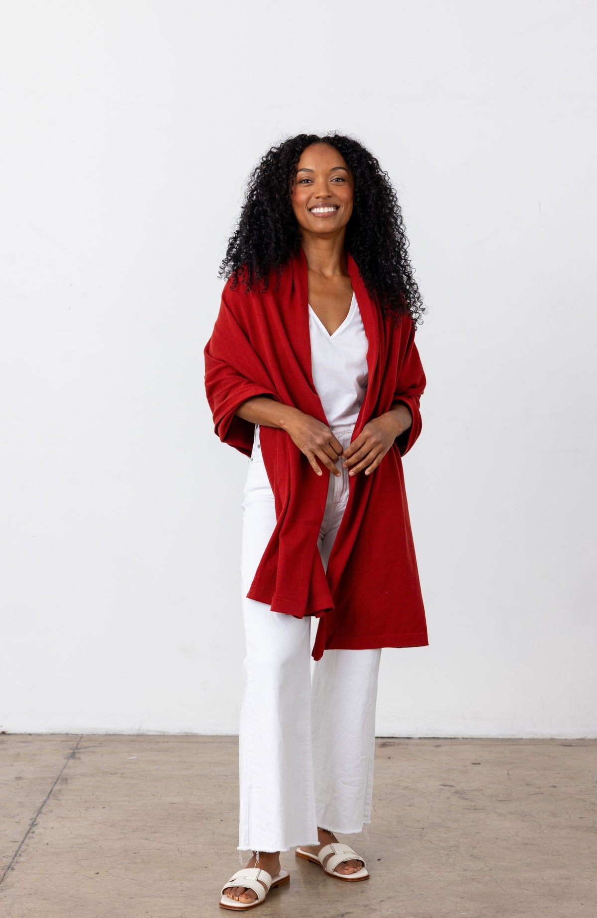 A woman with curly black hair stands against a white wall wearing a bright red draped cardigan over a white V-neck top and white wide-leg pants, with white slide sandals.