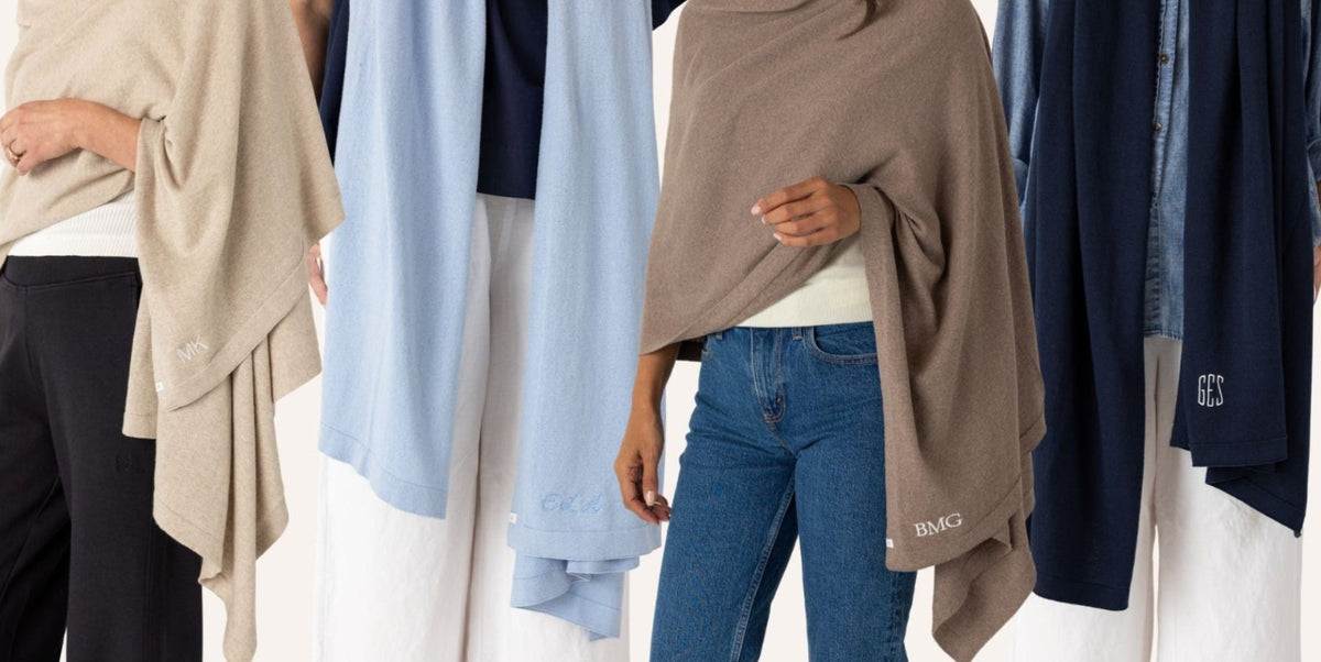A person wearing a taupe-brown shawl draped over their shoulders, paired with blue jeans and a white top, standing in front of a display of other shawls in light blue, navy, and beige.