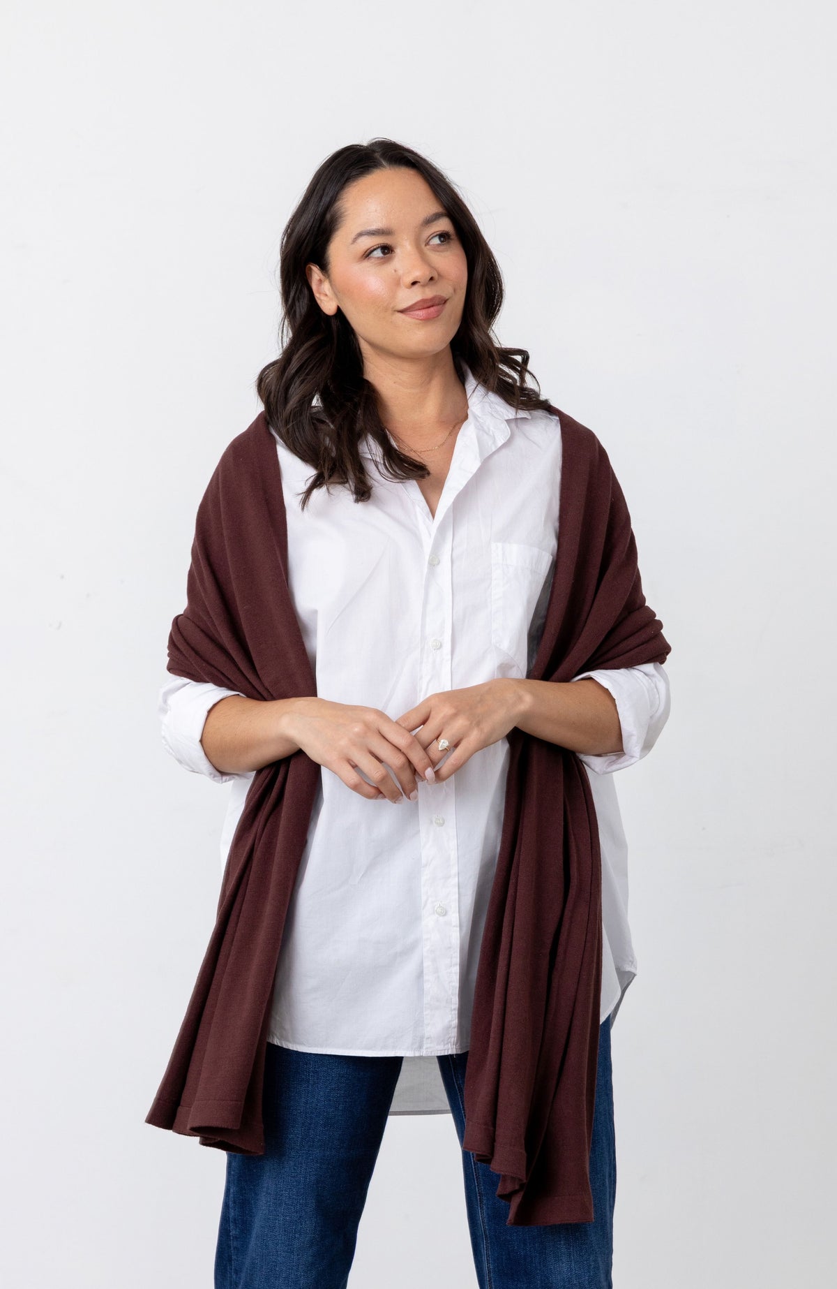 A woman modeling a dark brown shawl draped over her shoulders, worn over a white button-down shirt and blue jeans, against a plain white studio background.