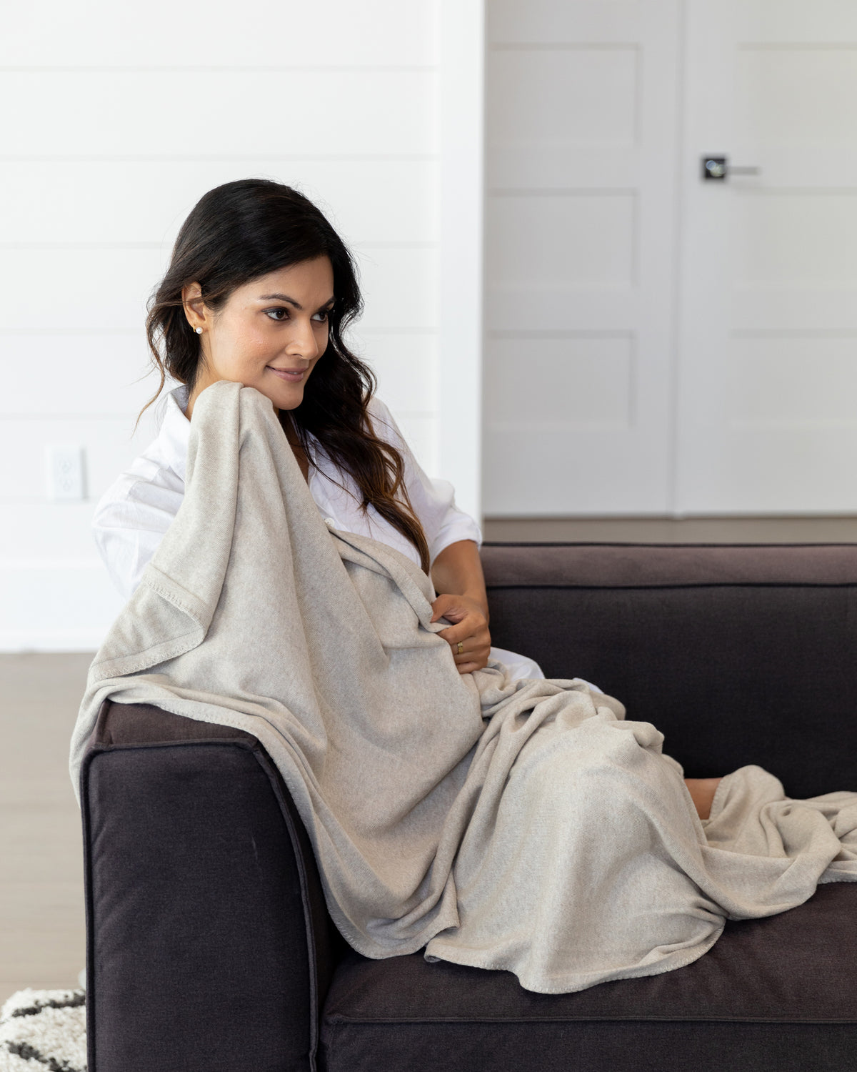 Woman sitting on the sofa using a Dreamsoft Travel Scarf in Birch Scarf which is a cream scarf as a blanket