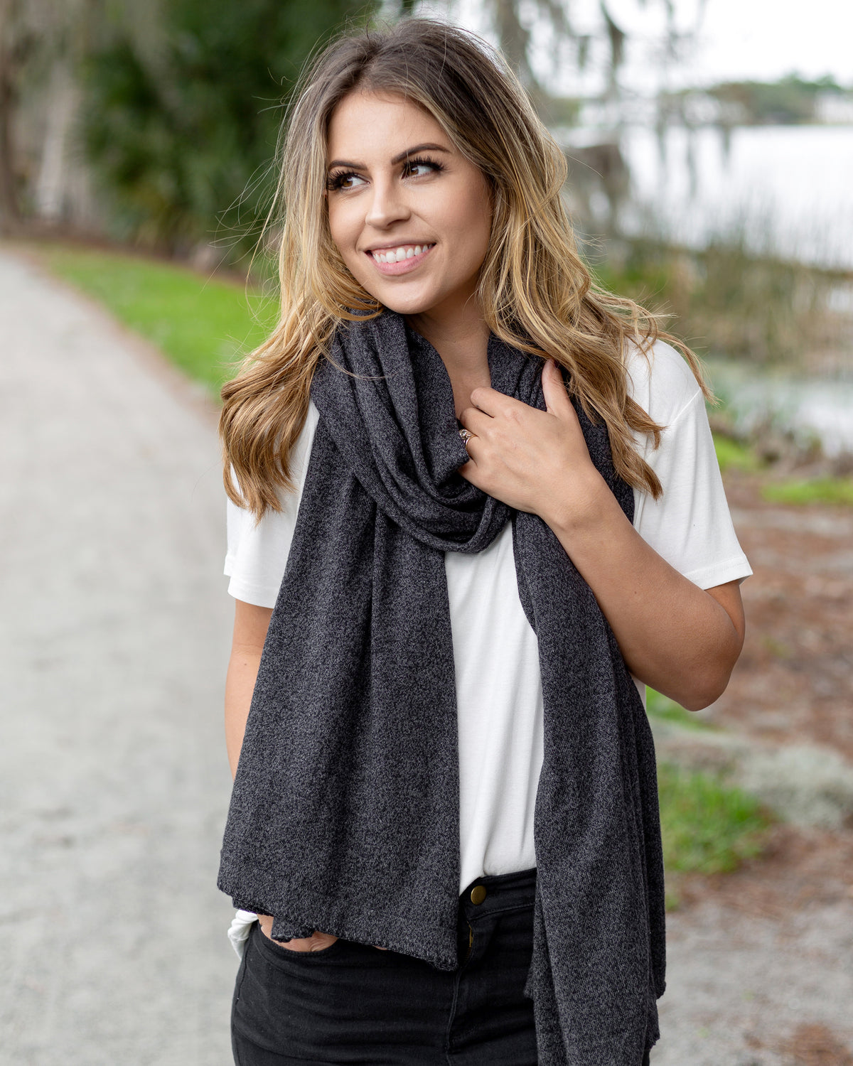 Woman wearing the Dreamsoft Travel Scarf in Graphite which is a gray scarf, worn as the modern loop with her hand in her pocket and other hand on the scarf