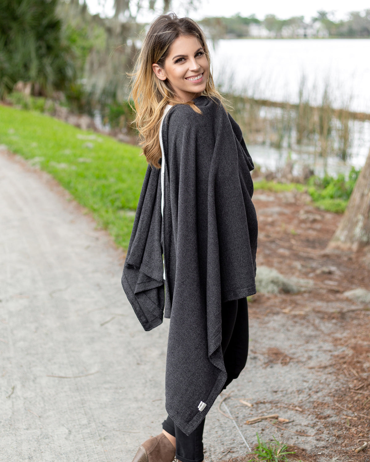 Woman wearing the Dreamsoft Travel Scarf in Graphite which is a gray scarf, worn as a blanket over the front of her body while standing on the side of a road
