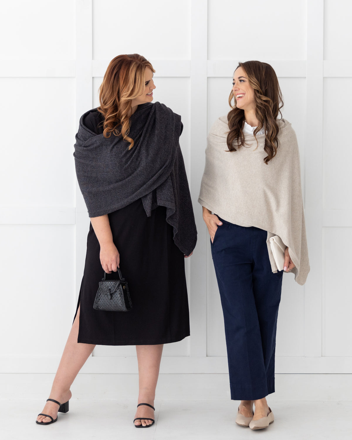 Woman wearing the Dreamsoft Travel Scarf in Graphite which is a gray scarf, worn as the wrap over her shoulder while holding a purse standing next to a woman wearing a Birch Dreamsoft Travel Scarf which is a tan scarf, looking at each other and smiling