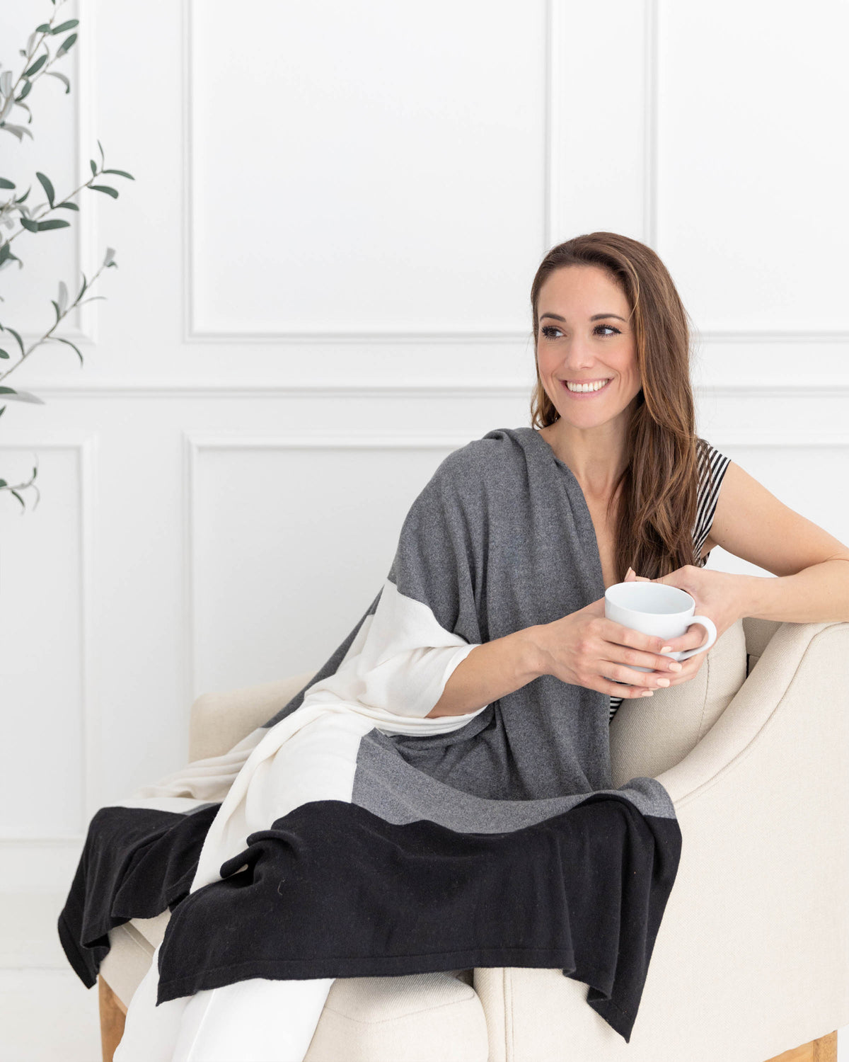 Woman wearing the Dreamsoft Travel Scarf in Gray Colorblock which is a black, gray and cream scarf, sitting on a chair while drinking coffee and smiling