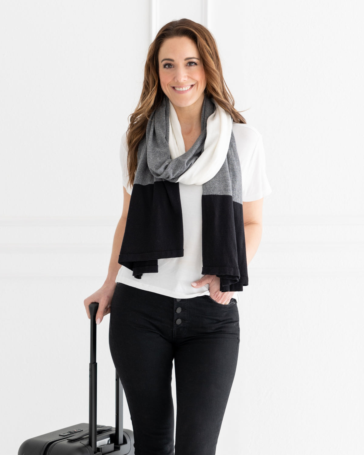 Woman wearing the Dreamsoft Travel Scarf in Gray Colorblock which is a black, gray and cream scarf, pulling a suitcase smiling at camera