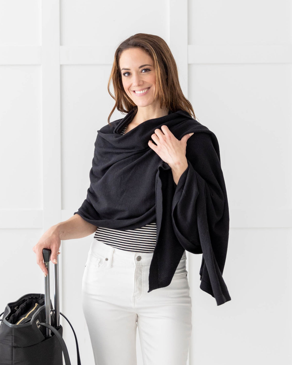 Woman wearing the Dreamsoft Travel Scarf in Black, wearing it as a wrap while  standing with her suitcase and bag